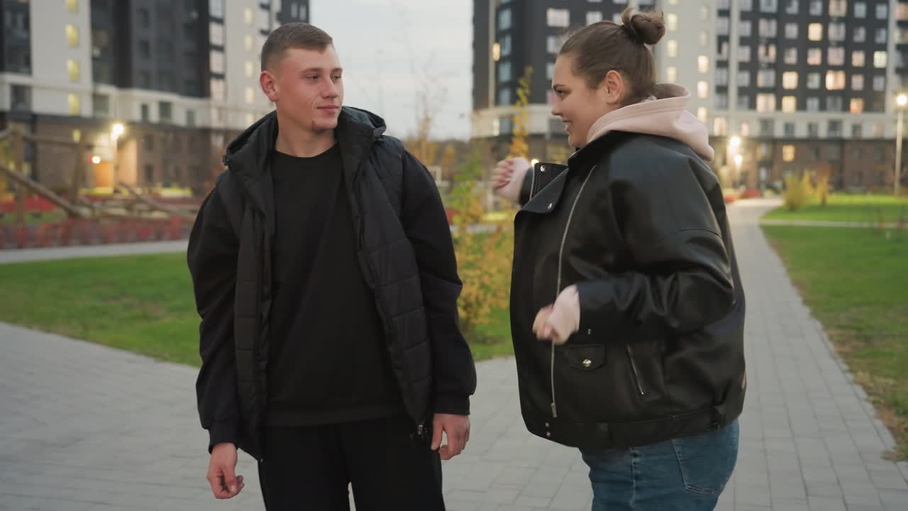 Funny moment as cheerful sister dances in front of her unimpressed brother during casual evening walk in urban park accidentally bumping into him while laughing and trying to lighten his mood
