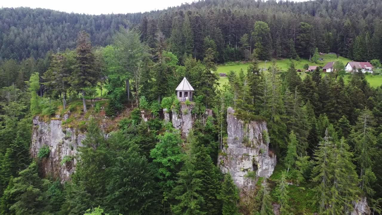 An aerial view captures a gazebo standing on a cliff, surrounded by a dense forest. Houses and mountain slopes are visible in the distance