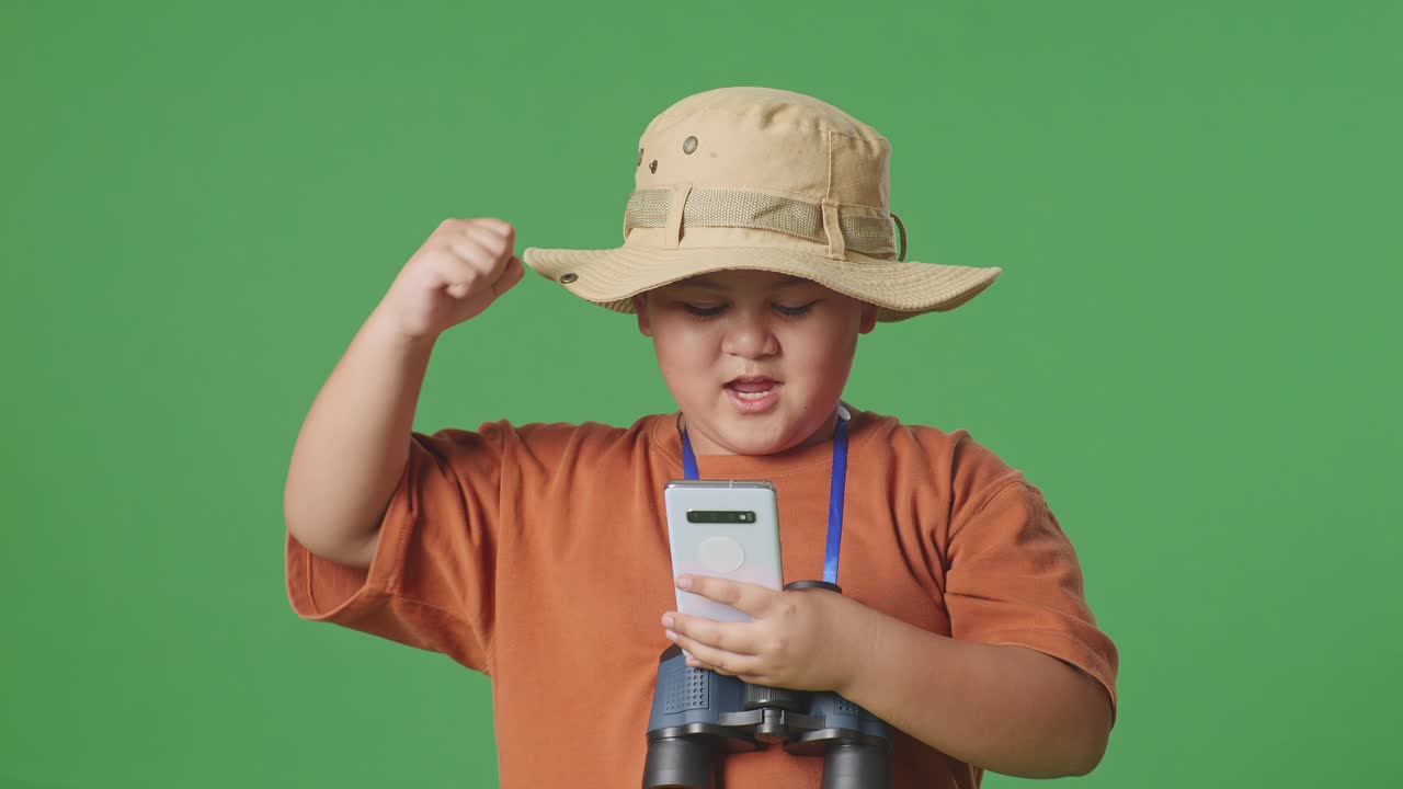 모자와 안경을 쓴 아시아 관광소년이 스마트폰을 보고 소리를 지르며 녹색 화면 배경에서 골을 축하하고 있다. 소년 연구원은 여행, 관광, 모험, 클로즈업