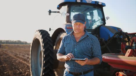 farmer is using digital tablet. on farm field, near tractor, on farm machinery backdrop. smart farming. farming technologies