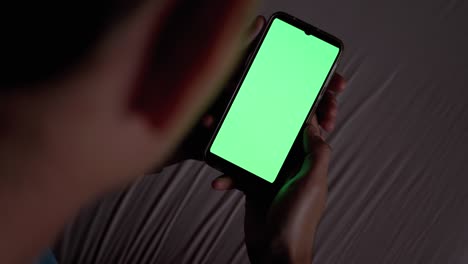 man in dark on bed holds and examines smartphone with green touchscreen