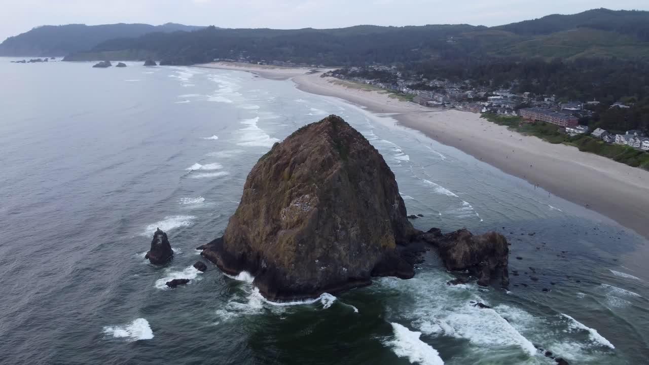 Waves rolling into Cannon Beach as the drone circles around Haystack Rock and birds fly around. Houses litter the beach as mountains rise above the forest in the background. 4K cinematic nature shot.