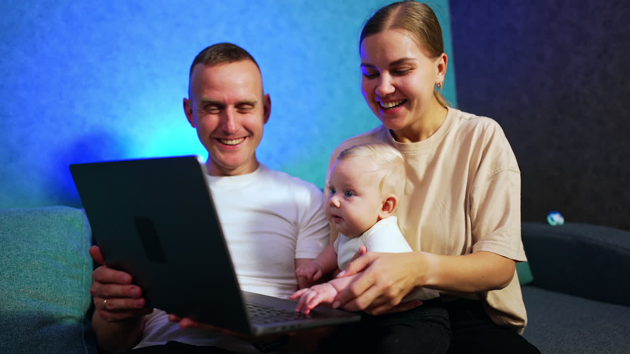 Happy Caucasian couple sitting close to each other. Woman is holding a cute baby and man is having a laptop in hands. Mom taps kid's hands and taps them by keyboard. Low angle view.