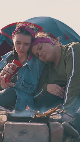 young lesbian couple with drink rests at burning bonfire near blue tent in calm camp on river bank at bright sunset