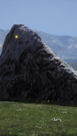 a large, grey rock sits in a grassy field with mountains in the background.