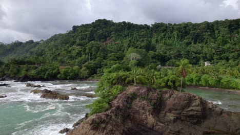 aerial dolly in of turquoise sea near rocky shore with green jungle hillside on overcast day, dominicalito beach, costa rica