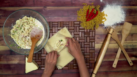 top view of rolling the spring rolls, marinated macaroni in the bowl, raw macaroni, wooden spoon, wooden spatula, flour, rolling pin, and chili on the table