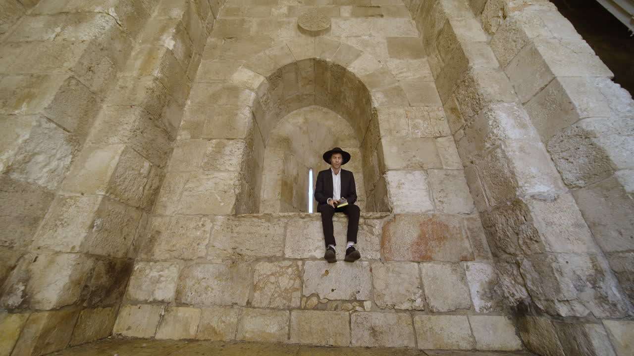 A man in traditional clothing sits in a stone archway, reading a book, amidst the ancient architecture of Jerusalem