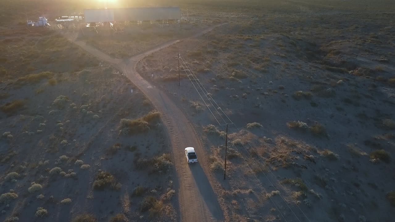 Vista aerea di un'auto fuoristrada su una strada sterrata su sabbia desertica, secca e rocciosa, con il paesaggio di lago e tramonto sullo sfondo.