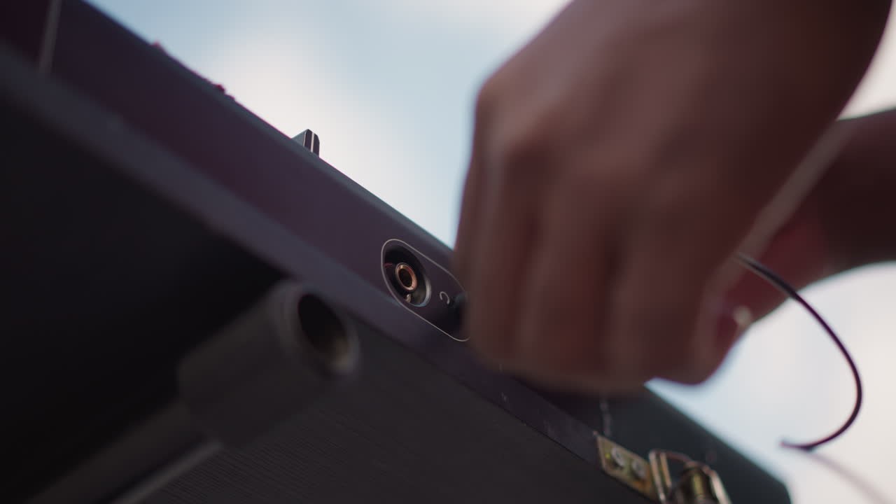 hands unplugging audio cable from speaker, street DJ packing gear after outdoor set, closeup of jack removal, dusk sky, empty stage and calm reflective mood, roadie tending flightcase