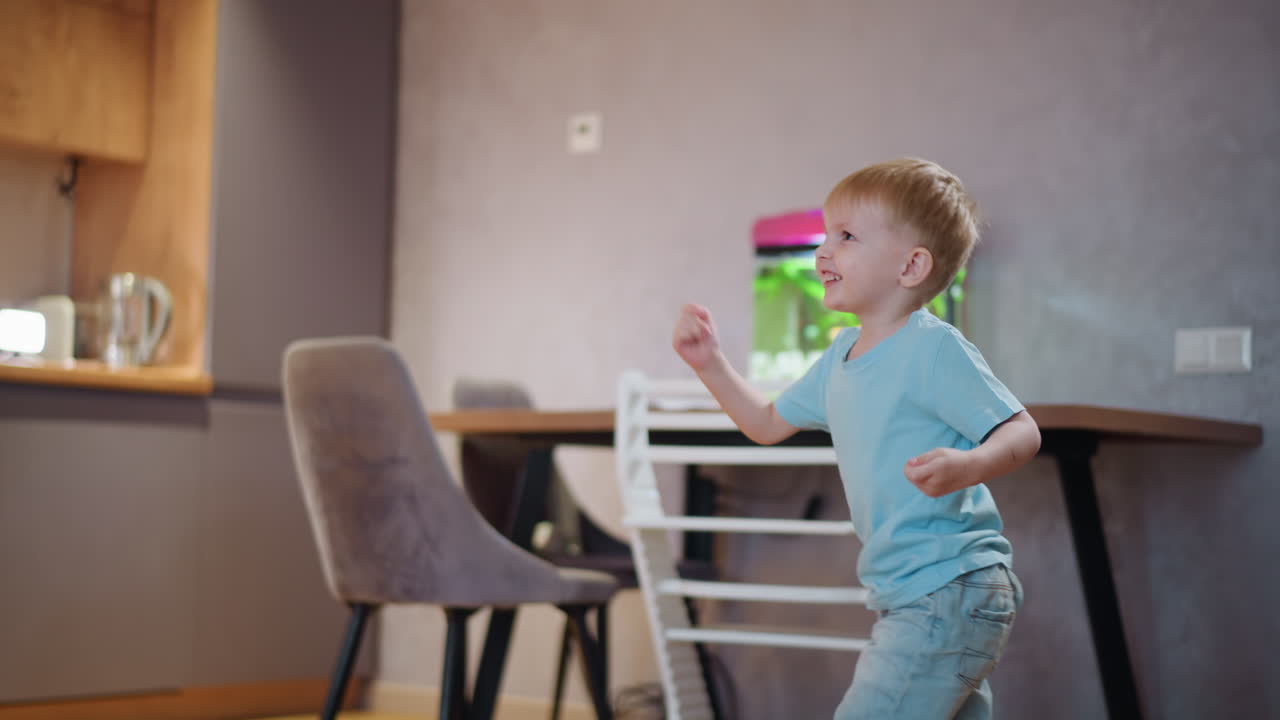 Happy young boy in blue shirt and shorts plays indoors near table with fish tank, smiling with excitement, enjoying carefree childhood fun, showing cheerful energy, joy movement inside cozy home