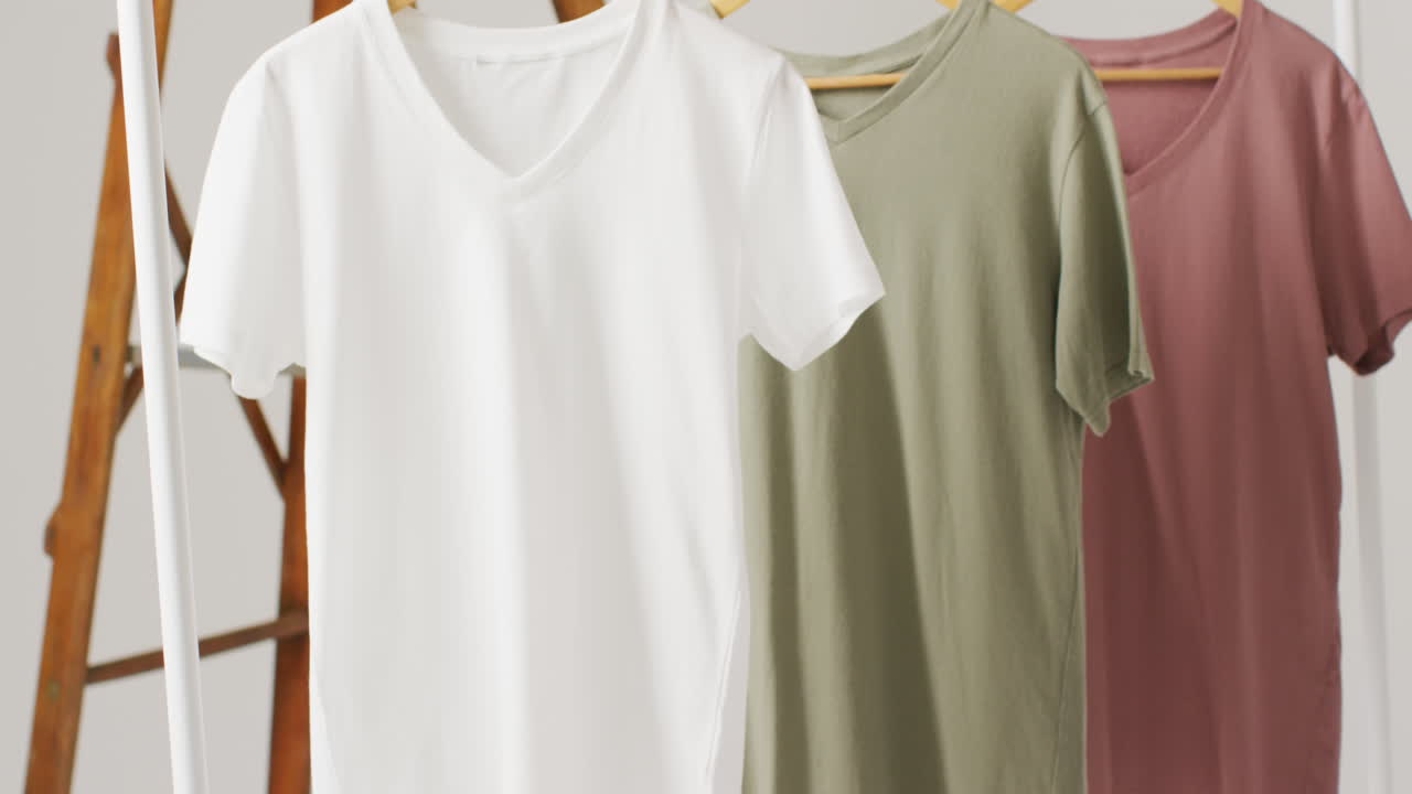 Video of close up of white, beige and gren t shirts hanging on white background