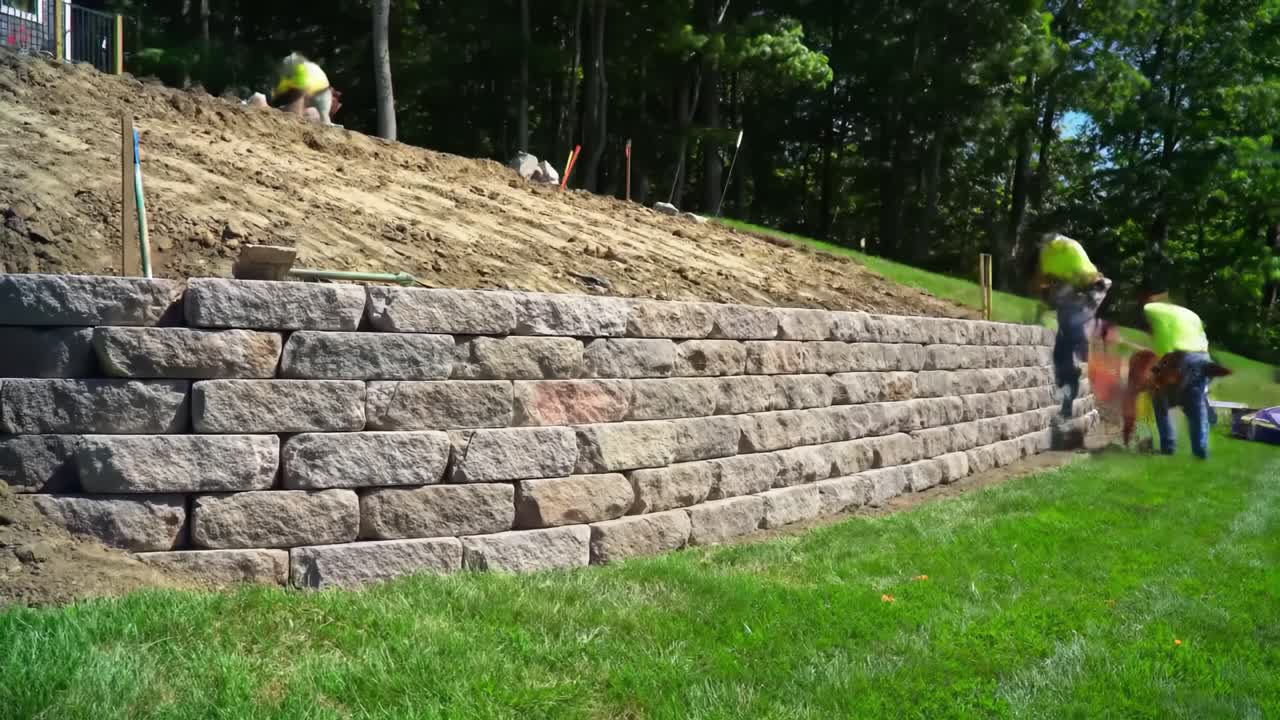 Skilled workers are building a resilient stone wall in a landscaped area surrounded by trees. The project showcases careful techniques and teamwork in outdoor construction.