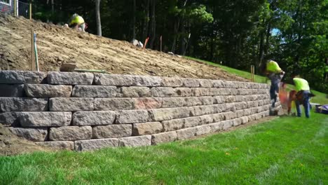 skilled workers are building a resilient stone wall in a landscaped area surrounded by trees. the project showcases careful techniques and teamwork in outdoor construction.