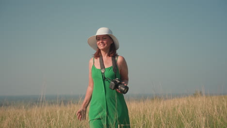 smiling mom in green sundress and white sunhat walking confidently through tall grass holding camera, enjoying peaceful outdoor landscape under soft blue sky, radiating joy