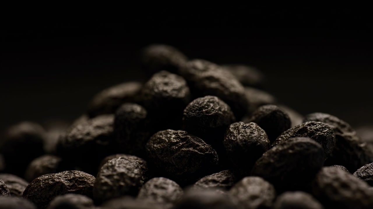 Aufgehäufte getrocknete Pfefferkörner oder ähnliche Gewürzbeeren liegen in einem niedrigen Haufen vor tiefschwarzem Hintergrund, mit scharfen Vordergrunddetails, groben, faltigen Schalen, subtilen Glanzlichtern und dunklem atmosphärischem Kontrast