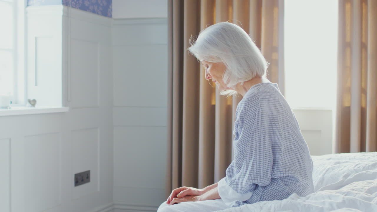 불행하고 우울한 노인 여성이 코로나19로 인한 봉쇄 기간 동안 집에서 침대 가장자리에 앉아 있습니다.