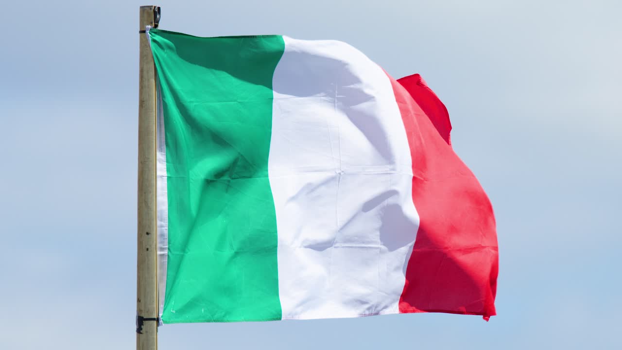 Italian flag flutters dynamically on flagpole under daylight, blue sky, steady camera, outdoor setting