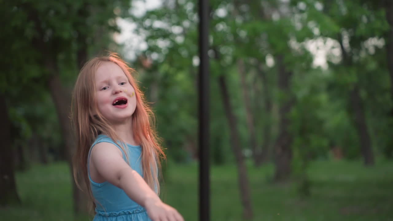 little girl chats while pushing friend on wooden chain swing in blurred green playground at dusk capturing playful interaction and joyful summer evening park fun among tall trees