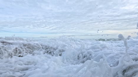 dynamic ocean waves crash against rocky shoreline under a cloudy sky, creating dramatic splashes and foam