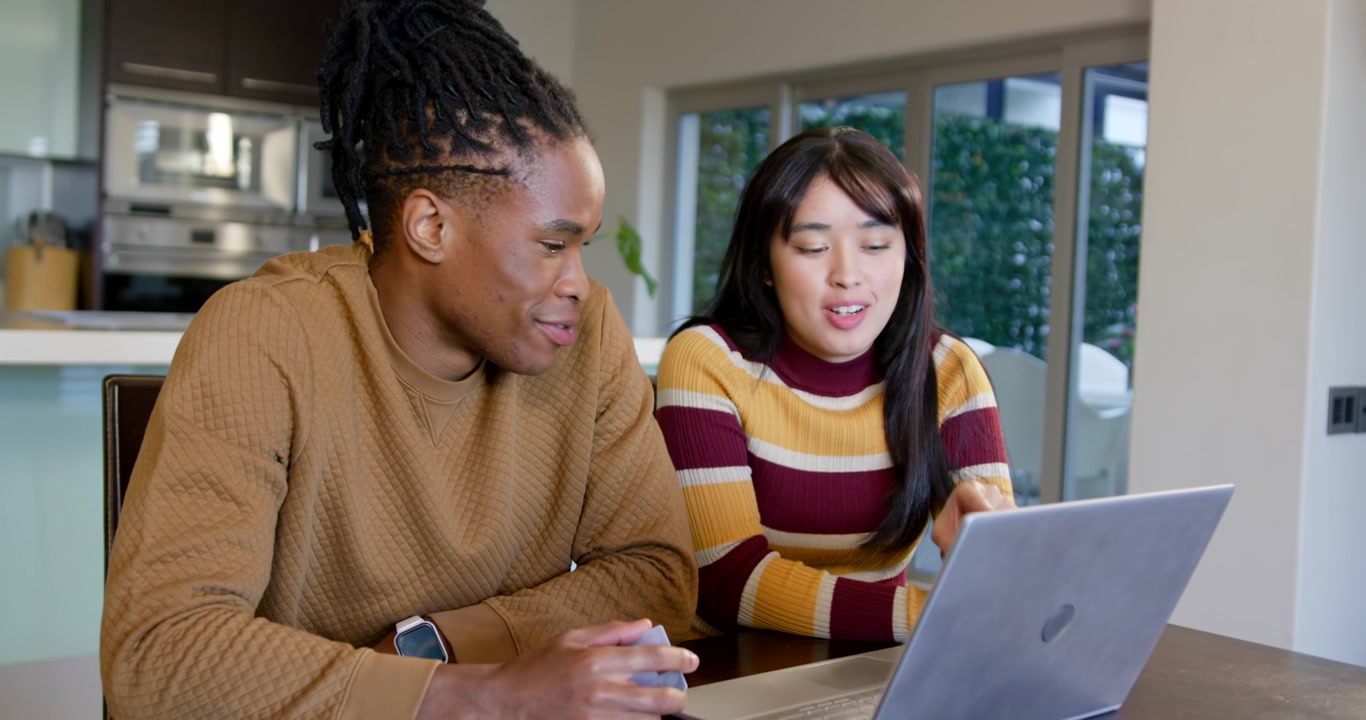 Diverse couple sitting at home table, man holding smartphone, gesturing, woman typing on laptop