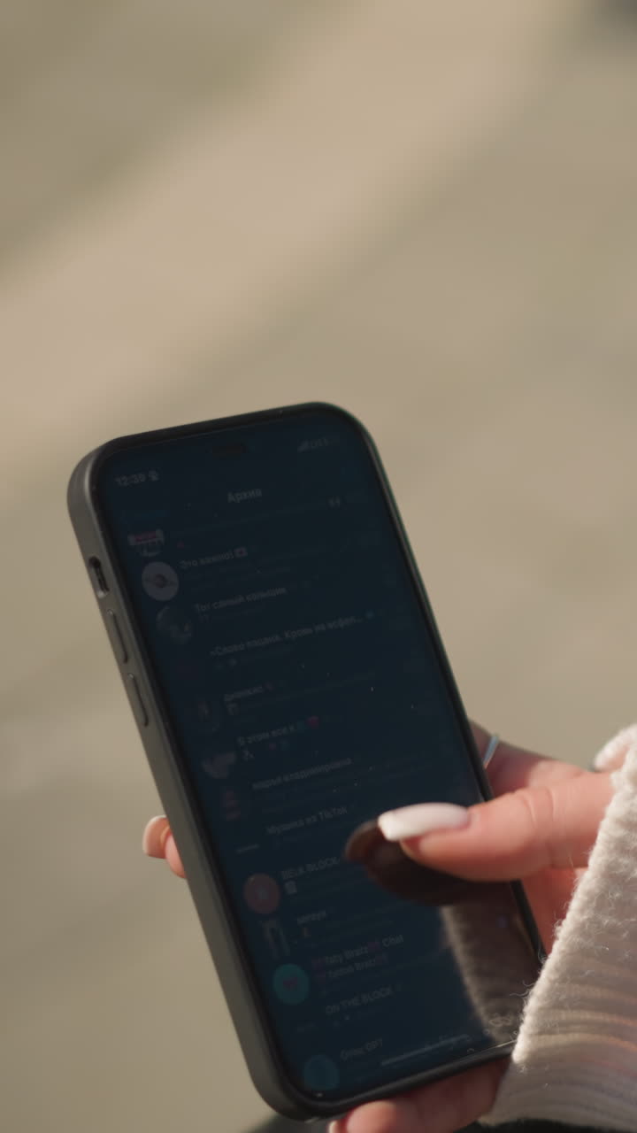primer plano de una mujer de cabello corto con abrigo negro y suéter blanco desplazándose en el teléfono inteligente con enfoque en la mano sosteniendo el teléfono mostrando manicure visible