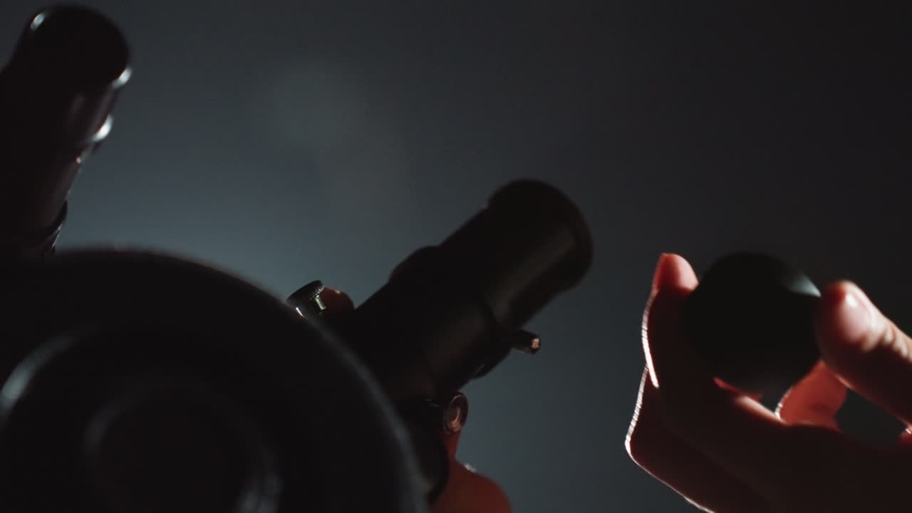 Hænder justerer teleskopets okular i svagt natlys, tæt intim indramning viser armbåndsur og fine fingre, der justerer fokus, dystert baggrundslys skaber silhuet, filmisk korn fremkalder