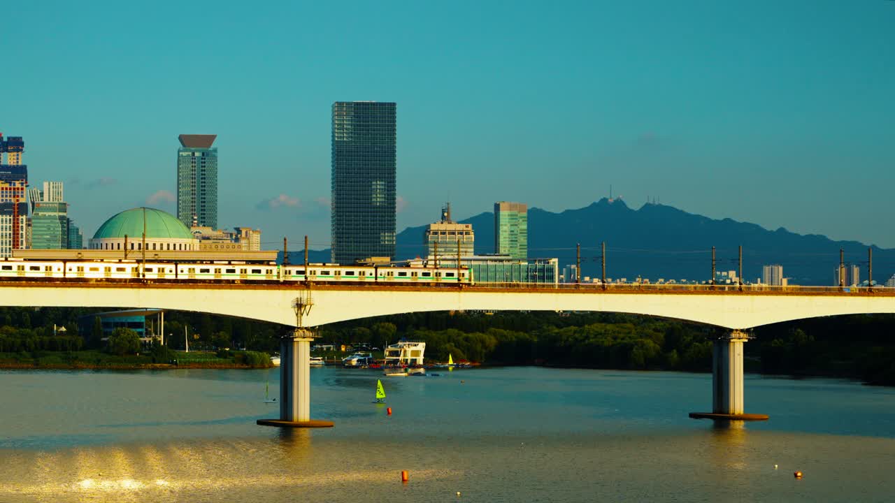 Seoul subway line 2 trains moving in opposite directions reflecting sunlight meeting on Dangsan Railway Bridge over Han River at sunset, National Assembly Building and Gwanaksan mountain in background