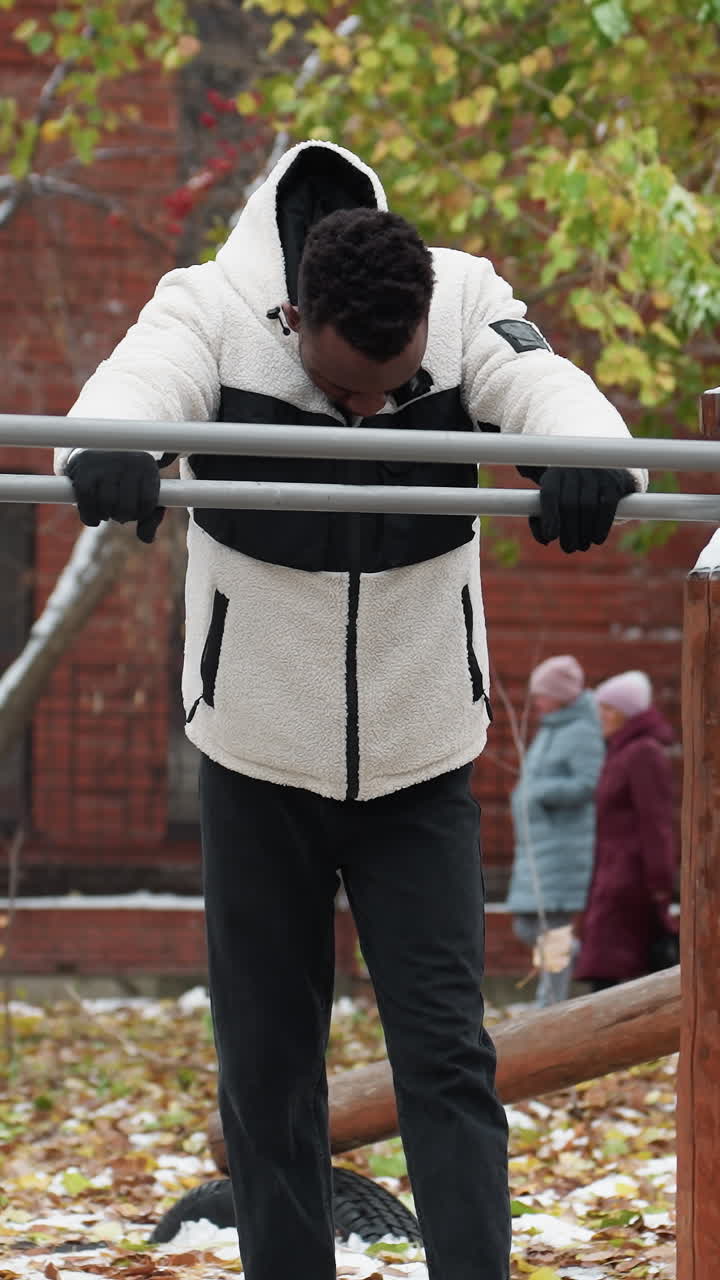 hombre negro descansando mientras sostiene una barra de hierro en el frente con la cabeza inclinada, vestido con una chaqueta de lana cálida y guantes, recuperándose de un entrenamiento intenso en invierno, personas trabajando en el fondo