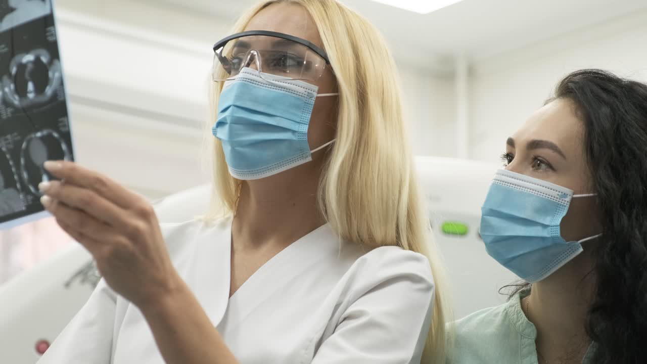 Woman doctor radiologist explains the results of CT scanning for young female patient, showing the snapshot with images, observing and analyzing CT scan in modern clinic beside modern CT scanner