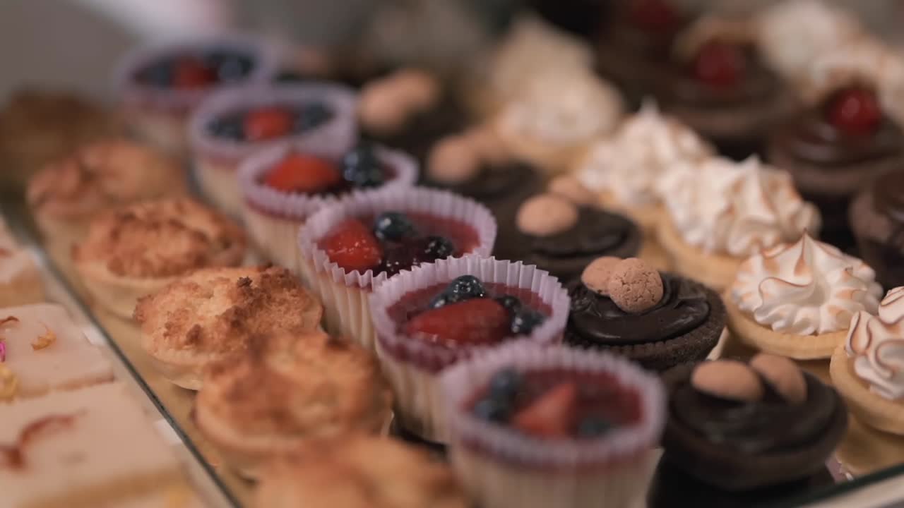 Primer plano con movimiento de barrido de una variedad de mini pasteles, que incluyen tartaletas de merengue de limón, dulces de chocolate y coberturas de frutos rojos, exhibidos en una elegante mesa de dulces para bodas