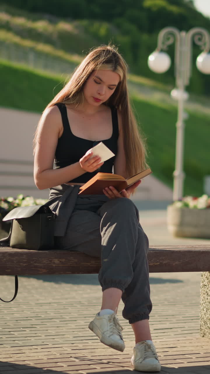 Woman seated outdoors on a bench with legs crossed, flipping a page from her book and observing a piece of paper thoughtfully, the background features lamp poles and a person seated in the distance