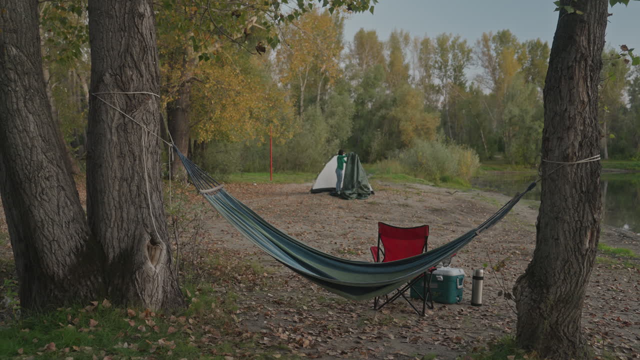 Træer indrammer hængekøje-campingplads i efterårsskæmring, rød stol og grønt telt i baggrunden, campingkomfur og udstyr i forgrunden, kølige aftentoner, forberedelse til nattur eller hvile i basecamp, rolig vildmarksstemning.