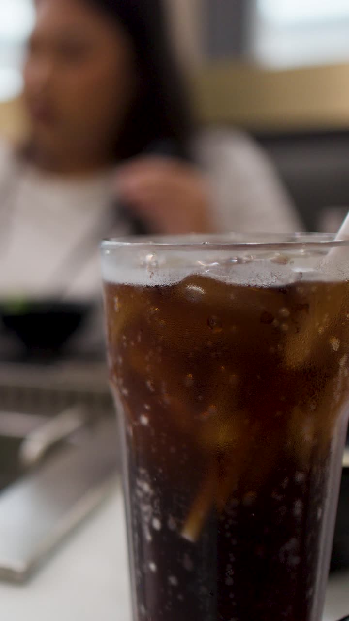 Primo piano di un bicchiere freddo di cola ghiacciata su un tavolo con una donna che cena sullo sfondo