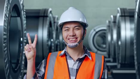 close up of asian male engineer with safety helmet smiling and showing peace gesture while standing in metal factory