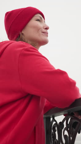 vista lateral de primer plano de una mujer con capucha roja y gorra de pie cerca de una barandilla decorativa de metal, mirando pensativa a la distancia brumosa, ambiente invernal sereno con iluminación suave y fondo borroso
