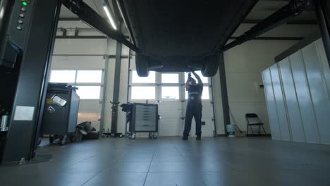 mecánico de automóviles con uniforme azul inspeccionando y reparando la parte inferior de un vehículo levantado en un taller de reparación profesional. el taller está bien iluminado y equipado con herramientas