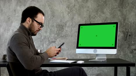 un hombre de negocios mirando sus mensajes de teléfono inteligente y sentado cerca de la pantalla de la computadora.