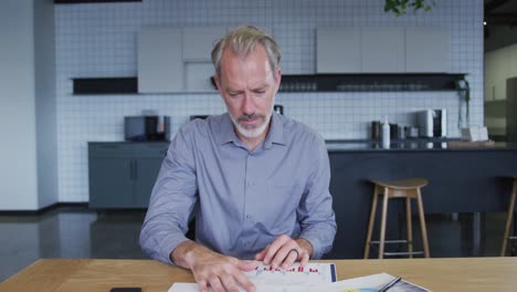 Senior-caucasian-businessman-having-video-chat-going-through-paperwork-in-workplce-kitchen