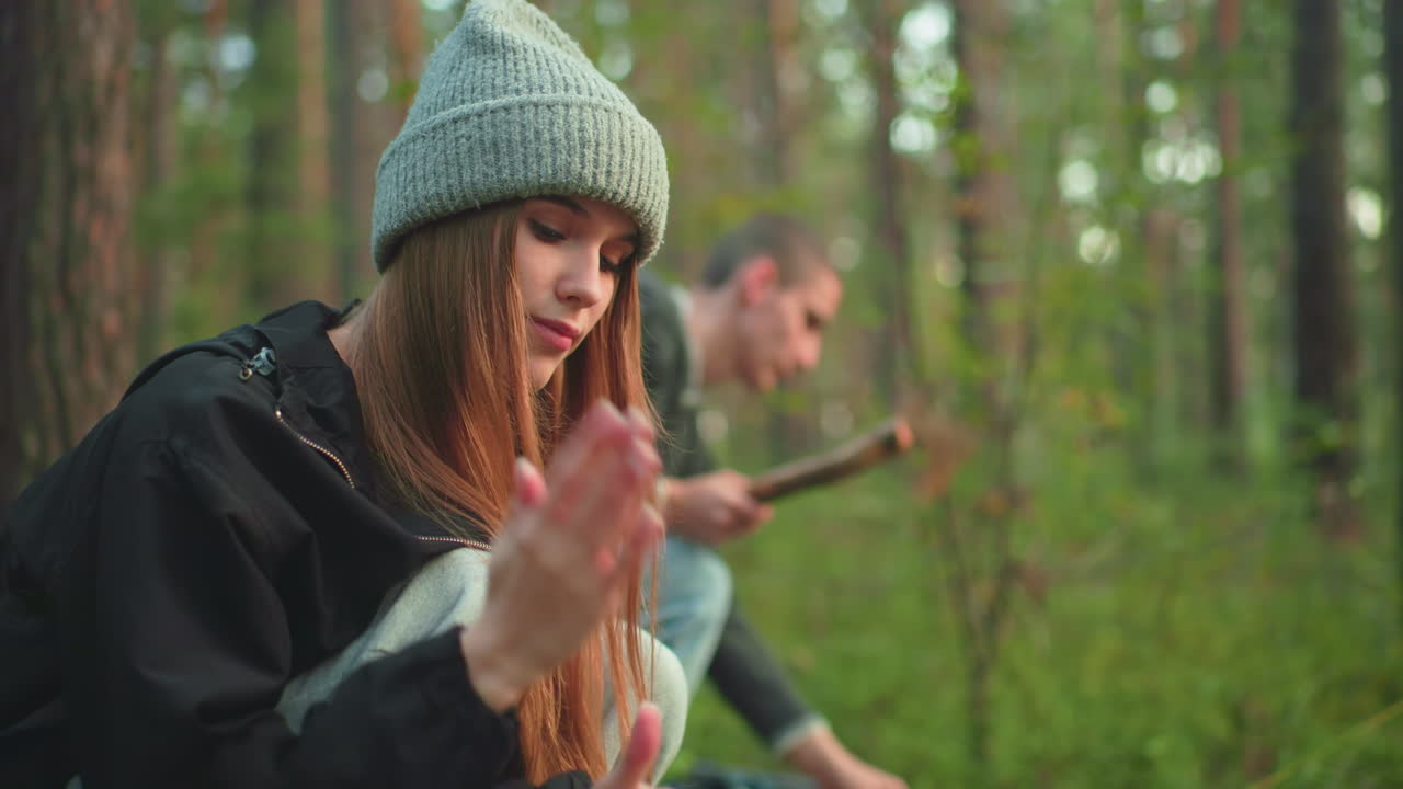 Young woman squatting in forest dusts hands after helping with tent setup while man beside her stands holding wooden stick preparing to adjust rope on tent during outdoor camping activity