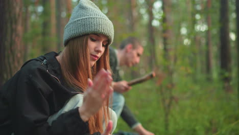 young woman squatting in forest dusts hands after helping with tent setup while man beside her stands holding wooden stick preparing to adjust rope on tent during outdoor camping activity