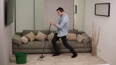 people, housework and housekeeping concept - happy man in headphones with mop cleaning floor and dancing at home. using swab pipe as a guitar. grey couch on the background