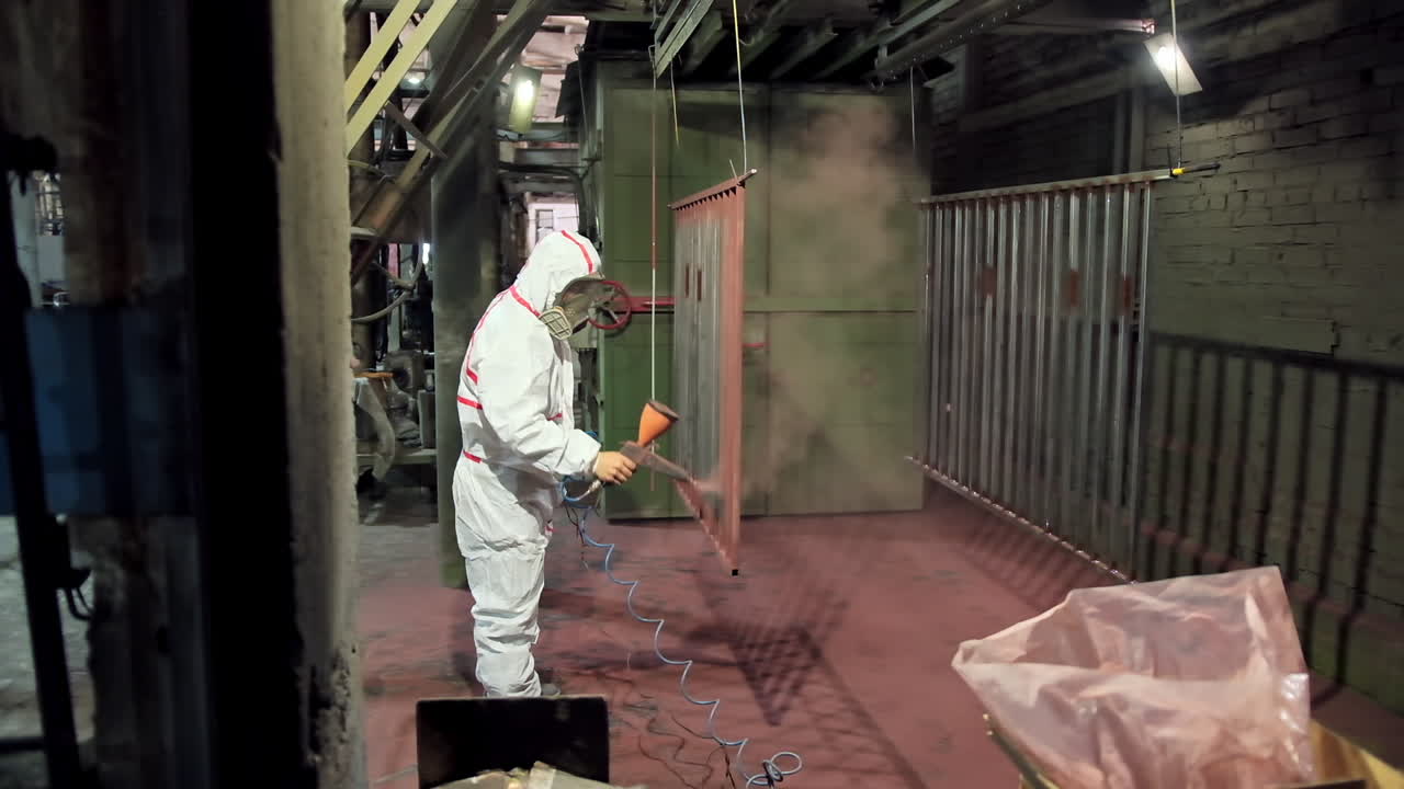 Painting parts in small factory production. Side view of a worker in protective suit working with a spray gun in a workshop. Man paints metal structure with a pulverizer.