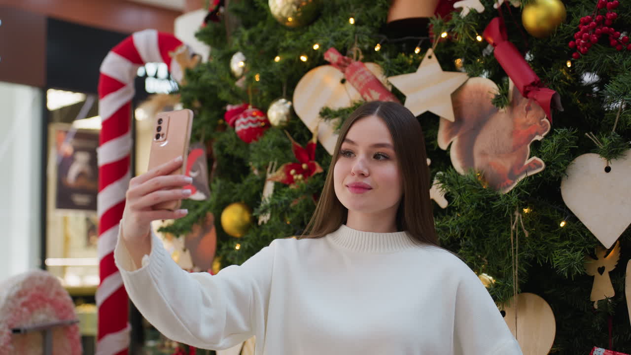 Young woman smiling and taking selfie with her phone in front of big decorated Christmas tree in shopping mall, capturing festive moment with colorful ornaments and holiday atmosphere