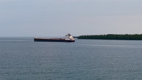 aerial drone view of a large cargo ship traveling across calm blue waters near a forested shoreline