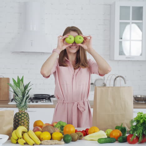 funny young woman closes her eyes with two green apples indoors. beautiful lady holds apples on her face and looks at camera in the light kitchen. healthy food concept.