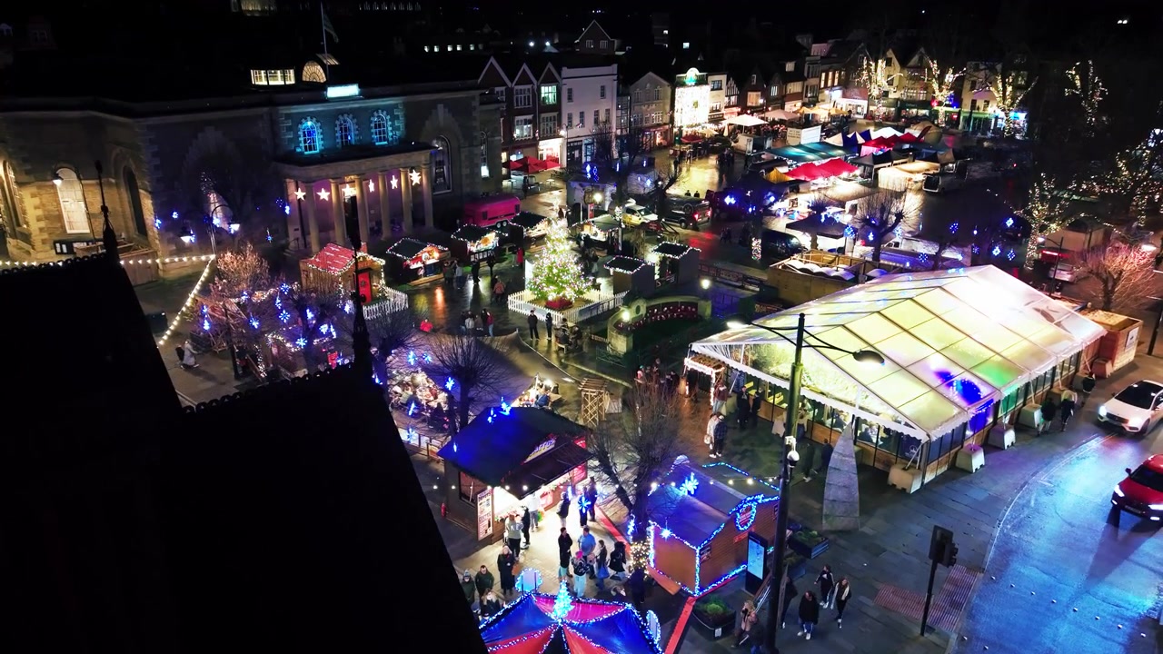 Flygfilm med drönare på natten över Salisburys julmarknad som visar en lång rad upplysta stånd, Guildhall och livliga folkmassor nedanför