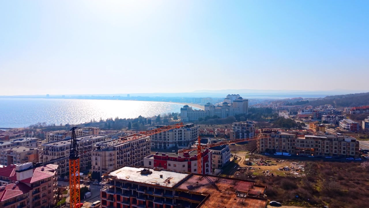 Cantiere edile costiero e località turistica. Ripresa aerea con drone di un edificio incompiuto con mare, spiaggia e hotel a cupola sullo sfondo.