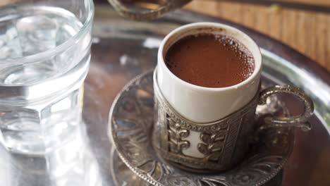 un primer plano de una mano sosteniendo una taza de café turco. la taza está en una bandeja de plata con un vaso de agua y una taza para café de plata.