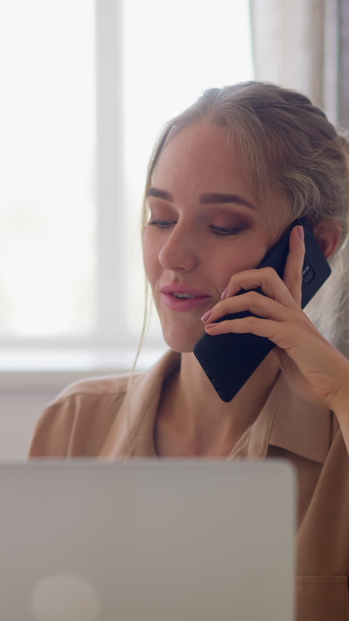 Young blonde woman talks on mobile phone sitting near of modern laptop at white table against bright window of office room slow motion